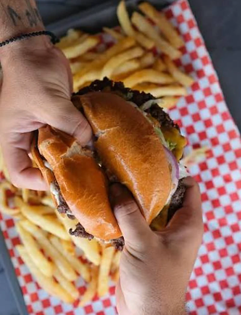 Two hands holding a smash burger over a tray of fries