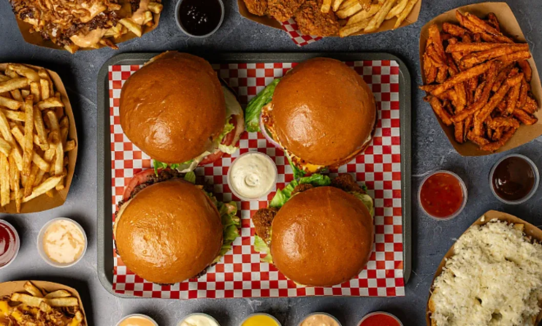 Four burgers on a red checked tray with fries, sweet potato fries and dipping sauces