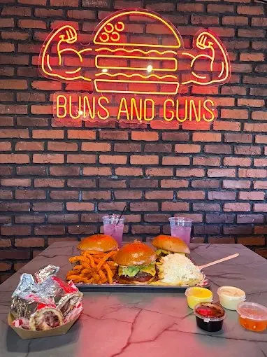 Three burgers, sweet potato fries and dipping sauces under the Buns and Guns neon sign